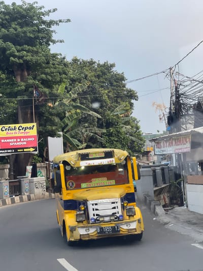 In South Tangerang, Indonesia, a yellow and blue public minivan, commonly known as an angkot, travels along a paved road with a slight curve. The vehicle has several advertisements on its exterior. One prominent sign, displayed on a yellow banner with red lettering, reads "Cetak Cepat Bisa Di Tunggu" and "BANNER & BALIHO SPANDUK". Another sign, a red banner with white lettering, advertises "AYAM AYAM". The angkot is adorned with various stickers and a phone number, "0812 9550 1267", and a price, "RP. 5.000", are visible. The license plate reads "B 1955 YJ".
The environment surrounding the road features dense foliage, including banana trees, and utility poles adorned with a tangle of electrical wires. Buildings with corrugated roofs and varying architectural styles line the opposite side of the street. The sky is overcast. The image captures a typical street scene in a bustling Indonesian city.