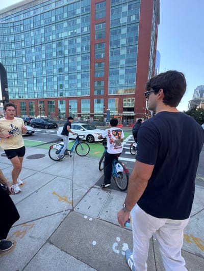 The image depicts an outdoor urban street scene in the Seaport District of Boston, United States. The background is dominated by One Marina Park Drive, a multi-story building featuring a red brick lower section and extensive upper levels constructed with a reflective glass curtain wall.
Five individuals are present in the foreground and midground. In the immediate right foreground, a male subject is viewed from behind, wearing a dark short-sleeved t-shirt, light-colored trousers, and dark sunglasses. He holds a small multi-colored object in his right hand. To his left, a partial view of another individual's lower legs and feet on the sidewalk is visible. Further left, a male subject wearing a yellow short-sleeved t-shirt with a graphic and black shorts is shown in motion, suggestive of jogging, with earphones in both ears. Two other male subjects are riding blue and white Bluebikes rental bicycles; one wears a dark t-shirt and light jeans, and the other wears a white graphic t-shirt with a print on the back and dark pants. Both cyclists are positioned on a green-painted bike lane adjacent to the concrete sidewalk.
The street behind the cyclists contains vehicular traffic, including a white sedan and a dark SUV. The concrete sidewalk features expansion joints and indistinct spray paint markings in yellow and white. Two metal utility access panels are embedded in the sidewalk. The sky above the buildings is clear blue.