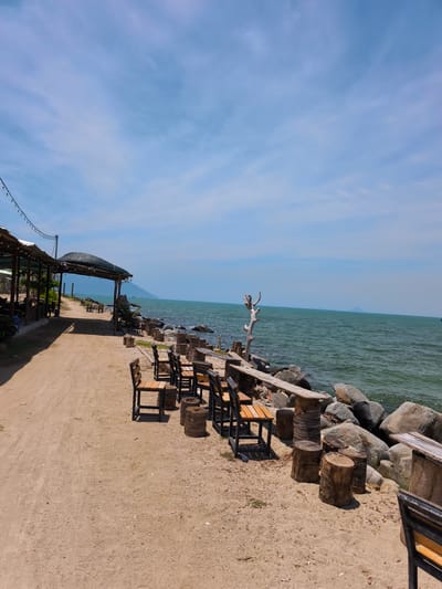 This is a photograph of an outdoor seating area situated on a sandy beach adjacent to the ocean in Vĩnh Lương, Vietnam. The scene is bathed in daylight under a partially cloudy sky. On the left side of the image, a covered walkway with a thatched roof runs parallel to the beach, supported by wooden posts. Several tables and chairs are visible beneath this structure, indicating a dining or lounging area. Further down the beach, more seating arrangements are present, including wooden chairs and tables made from tree stumps. The beach itself is composed of light-colored sand, with scattered debris and some vegetation. To the right of the seating area, large rocks border the ocean, and the turquoise water is calm with gentle ripples. In the distance, a hazy mountain range is visible on the horizon. The overall atmosphere is one of a relaxed, seaside establishment. There are no people visible in the frame.