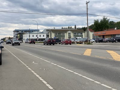 The image captures a street scene in Whitehorse, Canada, specifically looking east on Fourth Avenue near the intersection with Alexander Street. The weather appears overcast. On the left side of the road, several vehicles are parked or moving, including a black SUV, a dark sedan, and a red SUV. The road itself is asphalt with clear lane markings, including a dashed white line separating lanes and a solid yellow line on the far side.
Buildings line the north side of the street. The most prominent building in the foreground on the right is the Medical Dental Centre, an orange-paneled building with signage. Adjacent to it, to the west, is a pale green building with white trim, possibly an office or residential complex. Further west are multi-story buildings, one with a blue roof and modern architecture, and another smaller white building.
The background shows a densely forested hillside, characteristic of Whitehorse's natural surroundings. A wooden utility pole with electrical wires is visible on the right side of the image, and a street lamp stands near the center of the frame. The overall impression is of a commercial and residential area within a northern city.