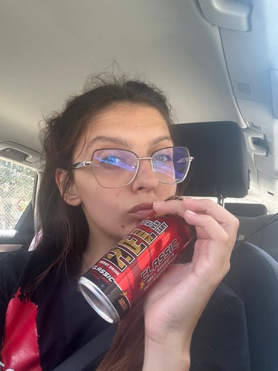 A young woman with long, dark hair and glasses is holding a can of energy drink in a car. She is wearing a black t-shirt with a red graphic. The can is red and gold with text that reads "Hell Energy Classic". The woman is looking at the can and appears to be about to drink from it. The background of the image shows the interior of a car, including the headrest of a seat. There is a window on the left side of the image, showing some greenery and a fence outside. The image appears to be taken during the daytime, with natural light coming in from the window. The location of Tryavna, Bulgaria is not visible in the image.