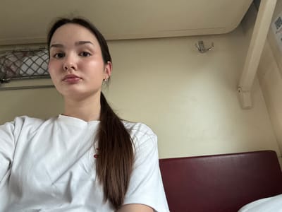 The image depicts a female individual within what appears to be a train compartment, consistent with typical Russian railway carriages, presumably en route to or from the city of Cheremkhovo, Russia.
**People:** One female, appearing to be a young adult with fair skin, dark brown hair styled in a ponytail, dark eyes, and full lips. She wears a plain light-colored (possibly white) crew-neck t-shirt with a small red detail on the left chest, and small stud earrings are visible in both ears. She is looking directly at the camera with a neutral expression.
**Objects and Environment:** The individual occupies the left and central foreground of the frame. The background reveals the interior of a confined space. To the upper left, a metal mesh luggage rack is present, containing a white, possibly textile, item. A silver-colored double coat hook is affixed to the light-colored wall paneling on the upper right. Below and to the right of the individual, a section of a dark red (maroon) upholstered seat or bunk is visible. The walls and ceiling are uniformly light-colored, consistent with interior paneling of a transport vehicle. A light-colored horizontal trim piece runs across the upper wall/ceiling junction, and a vertical structural element is present on the far right. Indiscernible blurred text or a graphic is partially visible on the far right wall panel. The overall lighting is even and suggests an interior, possibly artificial, light source.