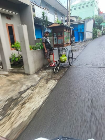 The image shows a man standing next to a small, wooden food cart with a single wheel and a green gas cylinder attached. The cart is parked on the side of a paved street in Cimenyan, Indonesia. The man is wearing a grey shirt, blue jeans, and a dark baseball cap. The street is wet and the buildings lining the street are residential, with a blue door visible. The man appears to be waiting for customers. The camera is located on a moving vehicle, with the street in the foreground. The image is slightly blurred due to the movement of the camera.