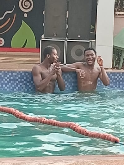 Two shirtless, dark-skinned men are waist-deep in a swimming pool with blue tiles. The man on the left holds his hands together while speaking, while the man on the right has his left hand on the pool's edge. A pool noodle separates the foreground. Large speakers sit behind the men, while a green leaf design adorns a wall in the background. A partial green chair is to the right.