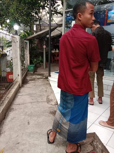 The scene takes place in the Kecamatan Ngaliyan district of Semarang, Indonesia, at the coordinates -6.992 latitude and 110.353 longitude. A man in a red shirt and blue sarong is standing on a concrete sidewalk, facing the camera. His left foot is on the ground, and his right foot is raised as if he is about to take a step. He has short dark hair and his expression is neutral. Behind him, to the right, is a second man wearing a blue denim shirt and brown pants. He is looking to his right and standing with his weight shifted to his left foot. In the background, a third person, whose legs can be seen, is standing on a tiled floor, looking down towards the sidewalk. The sidewalk is lined with a metal fence on the left side of the frame. The concrete surface of the sidewalk is cracked and uneven. The area behind the men is partially obscured by a leafy tree and the side of a building.  A small green trash can sits on the sidewalk near the fence. The tiled floor to the right of the sidewalk appears to be part of the entrance to a building.  There is an out-of-focus image of a car and what appears to be a street in the distance.  The day is partly cloudy and the lighting is bright.