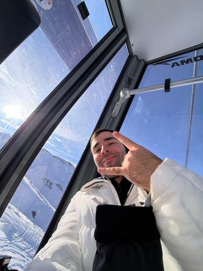 The scene depicts a male individual inside a ski lift gondola, situated within the mountainous region of Saint-Martin-de-Belleville, France. The man, appearing to be in his 20s or 30s with short dark hair and light stubble, is positioned in the foreground. He wears a white puffer jacket over a black high-collared garment. His right hand is raised, forming a "rock on" or "devil horns" gesture, with his thumb tucked in. He looks directly at the camera with a slight smile.
The gondola's interior features large, transparent glass panels that constitute the roof and sides, exhibiting visible surface scratches. These panels are framed by dark gray structural elements. A metal handle and a suspension cable are visible on the right side of the frame, integrated into the gondola's structure. Partial white lettering, "AMOM", is visible on a white ceiling panel.
Outside the gondola, a vast landscape of snow-covered mountains dominates the background under a clear, bright blue sky with scattered faint clouds. Sunlight is prominent from the upper left, casting bright reflections on the glass. Ski tracks are discernible on some of the snowy slopes.
