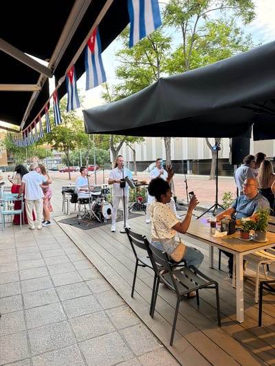 The image depicts a live music performance outdoors, likely in Cambrils, Spain, given the presence of Cuban flags. A band, composed of at least four members, plays on a raised platform. The band includes a drummer and a vocalist. Spectators are seated at tables near the performance area. A black awning partially covers the performance area. Several Cuban flags are strung along the awning's edge. A building facade is visible in the background. Some attendees are standing and watching the performance. The paving changes from sidewalk blocks to a boardwalk-like wood floor at the performance stage. One woman, seated, is taking a photo with her phone. The atmosphere appears relaxed and social. A group of people are standing by a table watching the show.