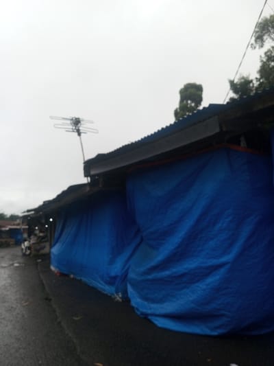 The image displays an outdoor scene in Sukanagalih, Indonesia, characterized by an overcast sky and a dark, wet ground surface, indicative of rain or recent precipitation. The left and mid-ground feature a row of market stalls or small commercial structures. The immediate foreground structures are largely covered by several large, royal blue tarpaulins, obscuring their contents. These structures possess dark corrugated metal roofs, with a metallic, Yagi-style antenna mounted on a pole extending from one of the rooflines in the mid-ground. Foliage from trees is visible above the rooflines against the gray sky. In the blurred distance to the left, a light-colored scooter or motorbike is parked next to a stall, and faint interior lights are discernible within some of the more distant structures. No individuals or direct interactions are clearly visible within the frame.