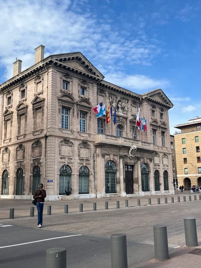 The image captures the façade of the Marseille City Hall, a prominent building in Marseille, France. The building's architecture is characterized by its ornate stonework, classical details, and arched windows with dark green grilles. Several French flags are displayed on the building. In front of the City Hall is a paved square with cylindrical bollards delineating a pedestrian area. A person wearing a brown jacket, blue jeans, and white shoes is standing in the square. Other buildings are visible in the background. The sky is a light blue with scattered clouds.