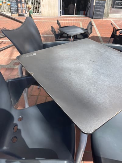The image shows a close-up view of a square, metal table with a smooth, grey surface, located on a brick-paved patio, in the heart of Las Palmas de Gran Canaria, Spain, at the given coordinates. The table is surrounded by black plastic chairs. Three of the chairs are empty, while the fourth is obscured by the table's surface. The table is in the center of the frame and is partially covered in shadow. The table's surface features a light, faded inscription, possibly a brand or logo. The patio is visible in the background, showing other tables and chairs, along with a building wall, and the faint outline of a figure walking away.