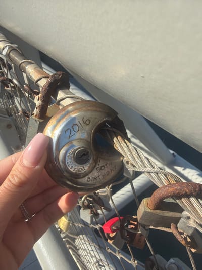 A close-up, high-angle photograph displays a light-skinned hand with manicured, light-pink fingernails holding a round, silver-colored stainless steel padlock. The padlock is engraved with "2016" across its top and "50TH BIRTHDAY" below the central keyhole. Its shackle is wrapped around a thick, braided metal cable. This cable is part of a larger metallic grid structure, to which numerous other padlocks of various sizes, colors, and states of corrosion are affixed, including some red and heavily rusted examples. A heavily rusted metal component is visible in the upper left of the frame. The background consists of a smooth, light-grey surface, potentially a wall or railing, above the padlock array, and a dark, indistinct area below, suggesting water or a significant drop. The scene is illuminated by direct sunlight. This structure is located in Cospicua, Malta.