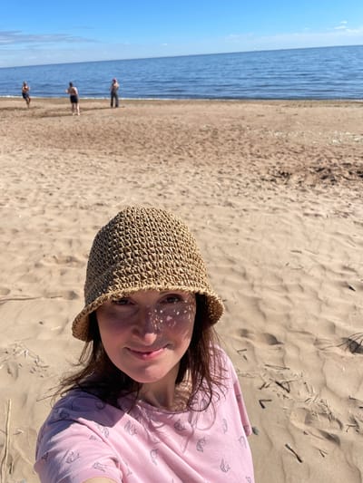 A woman wearing a straw hat and a pink t-shirt with a unicorn print is taking a selfie on a sandy beach. The sun casts shadows from the hat onto her face. In the background, the blue sea meets a clear sky, and three other individuals are visible in the distance. The beach is composed of sand with some scattered dry grass and twigs. The scene appears to be in Zelenaya Roshcha, Russia, on a sunny day.