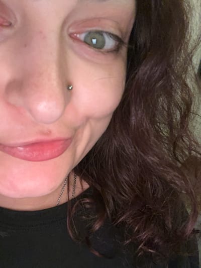 A close-up, informal portrait shot captures the left side of a person's face, taken from a slightly tilted angle. The primary subject is a person with fair skin, dark brown wavy or curly hair, and green/hazel eyes.

The left eye is prominently featured, open with a visible pupil and a small white reflection indicating a light source. The eyebrow is not fully visible, but some dark brow hairs are discernable. A subtle silver stud piercing is visible on the person's left nostril, suggesting a nose piercing. The lips are slightly parted, showcasing a natural pinkish hue, possibly enhanced with a lip product. The chin and jawline are partially visible on the lower right side of the image.

The person's hair, dark brown with some reddish undertones, fills the upper and right portions of the frame, appearing textured and slightly disheveled. A thin, silver-colored chain is visible around the neck area, partially hidden by the hair and ear. The background consists mainly of the person's hair and a dark, possibly black, fabric which could be clothing or bedding.

The lighting appears soft and even, highlighting the facial features without harsh shadows. The expression on the visible part of the face is neutral and calm. No specific activities are taking place beyond posing for the photo. The image provides no specific visual cues to its location in Picassent, Spain, or the time of day. There is no visible text.