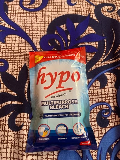 A packet of "Hypo" brand multipurpose bleach is placed on a patterned fabric. The fabric has a repeating pattern of brown and white squares with blue ornate swirls. The bleach packet is predominantly light blue with red and white text. Prominently displayed on the front are the words "hypo" in large red script, followed by "Go Wipe O!" and "MULTIPURPOSE BLEACH". A red strip at the top reads "NEW PACK KILLS 99.9% GERMS". Below the main text are three circular icons. The left icon depicts a gloved hand holding a blue cloth. The middle icon shows a folded white garment. The right icon illustrates a stylized representation of a white cloth with blue arrows emanating from it. At the bottom right corner of the packet, a red rectangle contains "75ml". The image is taken from a slightly elevated angle, focusing on the product. There are no people visible in the image. The environment is the patterned fabric itself, which appears to be a tablecloth or similar textile. The location is Kakau, Nigeria.