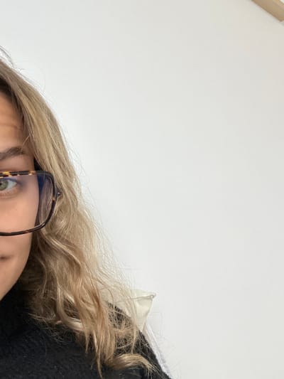 The image displays a partial, close-up view of an individual in an indoor setting within Veauchette, France. The subject, occupying the left third of the frame, is a person with wavy, light-colored hair, appearing blonde or light brown. The visible portion of the face includes the left eye with a green iris and the left eyebrow. The individual is wearing eyeglasses with tortoiseshell-patterned frames, the left lens and frame prominently visible. A dark, textured garment, resembling a sweater, is visible on the subject's lower left, with a white collar or undershirt peeking out near the right shoulder area. The background is a uniformly white, unadorned wall, filling the majority of the right and upper portions of the image. No specific actions or interactions are depicted; the individual appears stationary.
