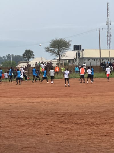 The image depicts a soccer match taking place on a dirt field in Du, Nigeria. Numerous individuals are present, both on the field playing and off the field as spectators. The players are wearing various colored jerseys, including blue, white, and orange. The field itself is predominantly red-brown dirt with some patches of grass. In the background, there are several buildings, a water tower, a communication tower, and trees. The sky is overcast. The overall scene suggests a casual sporting event in an outdoor urban or semi-urban setting within Du, Nigeria.