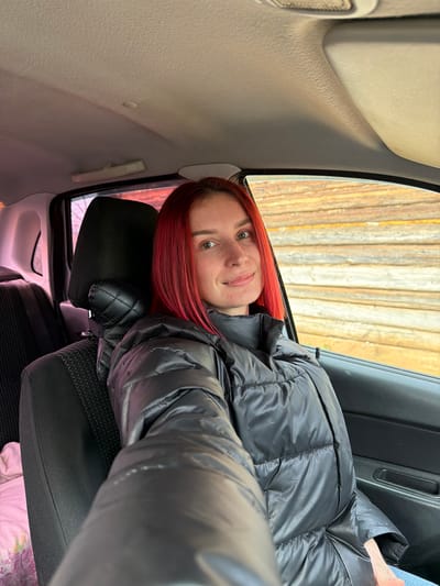 A woman with red hair wearing a black puffer jacket sits in the back seat of a car. Outside the car window, vertical wooden boards are visible. The location is Заболотово, Russia.