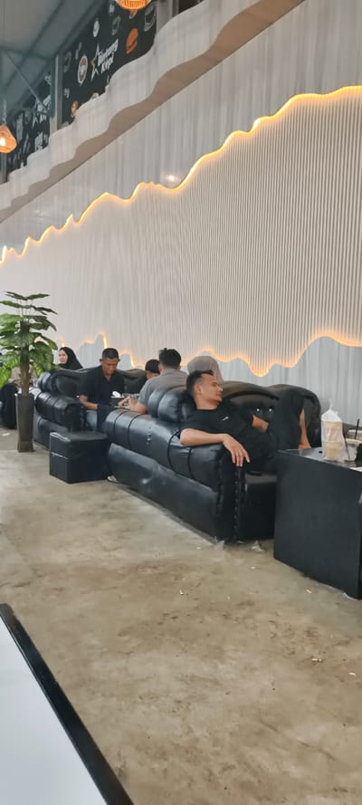 An indoor scene within a cafe or lounge in Matangjareung, Indonesia, features multiple individuals seated on black, modular faux-leather sofas. The environment includes a light-colored concrete floor with scattered debris, and a decorative wall composed of white vertical slat panels. This wall has a wavy top edge illuminated by a warm-toned LED strip light. Above this, a darker wall section displays black and white illustrations with orange accents and text reading "Bintang Kopi." The ceiling is high and industrial in appearance.

In the foreground, a male with short dark hair, dressed in a black t-shirt and black pants, reclines on a black sofa, his bare feet resting on the cushion. He looks towards the right side of the frame. Adjacent to him is a small black square table holding a plastic cup with a beverage and a white plastic bag. Behind him, another male in a dark shirt is seated, looking down at an object in his lap. Further left, an artificial green plant in a dark planter separates seating areas. In the background, several other individuals, including a person in a dark hijab, are seated on similar sofas. Most individuals appear to be relaxing or engaged with personal devices.