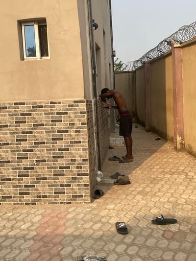 This image depicts an outdoor alleyway or passage between two buildings in Eziobodo, Nigeria, under clear daytime conditions. The ground is paved with hexagonal interlocking blocks in shades of tan and light brown. On the left, a light tan-colored building features a lower section clad with multi-toned brick-patterned tiles and a rectangular window with reflective glass and an aluminum frame on its upper facade. A dark outdoor light fixture is mounted on this building's wall. On the right, a plain tan/orange wall is topped with coils of razor wire.
A single male individual, bare-chested and wearing dark shorts with a visible red waistband pattern, is positioned on the left side of the passage. He leans forward, pressing his forehead against the light tan wall. His right hand holds a small, dark object, possibly a mobile phone, against the wall near his head. His left hand is also near his head. He wears dark slide-style sandals.
Scattered on the paved ground in the foreground and near the individual are several pairs of dark footwear, including black Crocs-style clogs and multiple black slide sandals, one distinctly marked "CARTER" and another with a white logo on its strap. Shadows cast by the buildings indicate sunlight from a high angle.
