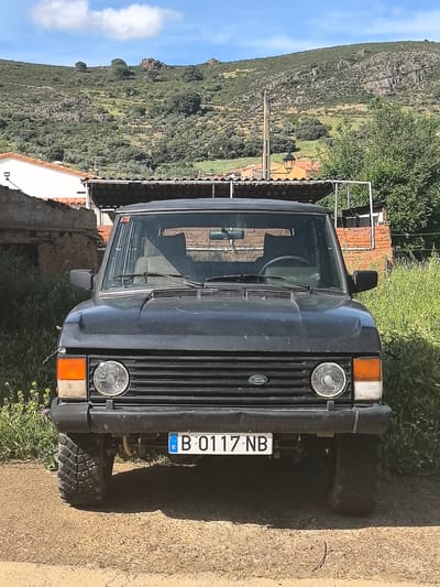 A dark-colored, older model Land Rover is parked on a dirt surface in what appears to be a rural setting. The vehicle faces the camera, showcasing its front grill, headlights, and license plate (B 0117 NB). The background features a landscape with a building, a hillside covered in low vegetation, and a blue sky with clouds. The building appears to be an old structure with a tiled roof. The location is likely Fontanarejo, Spain.