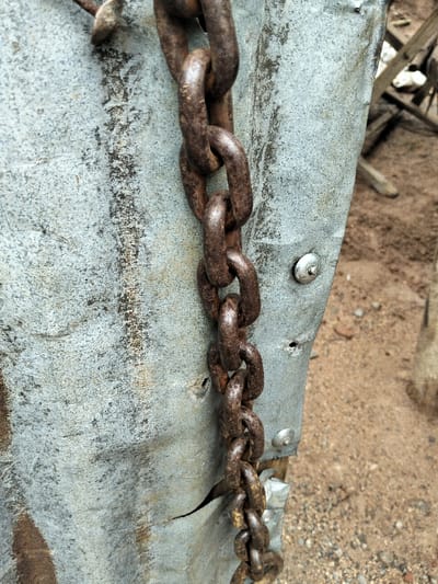 This image depicts a heavy, oxidized metal chain hanging vertically against a weathered, corrugated galvanized metal sheet in Nyansiongo ward, Kenya. The chain, dark brown with extensive rust, consists of thick, oval links. The light gray metal sheet exhibits significant dirt accumulation, dark rust streaks, and surface corrosion. Several metallic rivets or screws are visible on the sheet, securing it. The sheet itself shows signs of damage, including small holes and a prominent vertical tear towards the bottom. The background to the right reveals light brown, sandy ground with scattered small pebbles. No individuals are present in the scene.