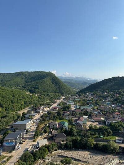 The image presents an aerial view of a settlement nestled within a valley, surrounded by lush green mountains. The settlement appears to be a town or village, characterized by numerous residential buildings with varied roof colors, including red, green, and brown. A paved road runs through the heart of the settlement, lined with houses and some commercial buildings. Several vehicles, including cars and vans, are visible on the road and parked near buildings.
A prominent feature is a multi-story modern building with a gray facade and a dark roof on the left side of the image, featuring a large banner with text and graphics. Adjacent to it, another modern building with a dark roof is visible. Further down the road, there are more traditional-looking houses and smaller commercial structures.
The natural environment is dominant, with steep, densely forested mountains on either side of the valley. A riverbed, filled with rocks and some flowing water, runs along the lower edge of the settlement. The sky is clear blue with a few wispy clouds. The overall scene depicts a quiet, picturesque community integrated with its mountainous surroundings.