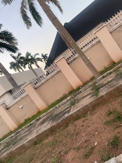 The image shows a street view of a building in Wauro Jabbe, Nigeria. The building has a beige exterior with a dark tiled roof and a white decorative fence. Palm trees are visible in the foreground and background. A concrete pathway runs alongside the building, leading to a dirt and gravel ground. A utility pole stands near the building, with some markings on it. The sky is overcast.