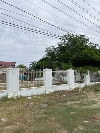 The image captures an outdoor scene in Bundar, Indonesia, featuring a white metal fence bordering a property. Behind the fence, lush green trees and a palm tree are visible, suggesting a tropical environment. The sky is overcast with clouds, indicating it might be a cloudy day. In front of the fence, the ground is covered with dry grass and scattered debris, including plastic bags and a red object that could be a piece of packaging. A portion of a building with a reddish-brown roof is also visible to the left. Several power lines crisscross the sky, dominating the upper left portion of the frame. The overall impression is one of an outdoor, possibly residential or commercial area, with a mix of natural elements and urban infrastructure. No specific activity or event is clearly depicted, and the scene appears static. The weather seems overcast, and the location cues point to a tropical setting. The image is taken from a low angle, looking upwards towards the sky and the fence. Some text is visible on a green sign behind the fence, but it is illegible due to the resolution and angle.