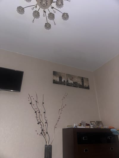The image depicts an indoor scene, likely a bedroom, with a neutral-toned wall that has a subtle sparkling texture. In the upper part of the image, a modern chandelier with clear crystal elements hangs from the ceiling. To the left, a flat-screen television is mounted on the wall. To the right of the television, a triptych canvas print featuring a London cityscape with Big Ben is displayed. In the foreground, a tall, dark vase contains a decorative arrangement of thin branches with small red and black beads. Below the canvas print and to the right, a dark brown dresser is visible, topped with various items including picture frames, bottles, and folded fabrics. The lighting appears to be dim. There are no people visible in the image, and no actions or interactions are taking place. The scene is static.