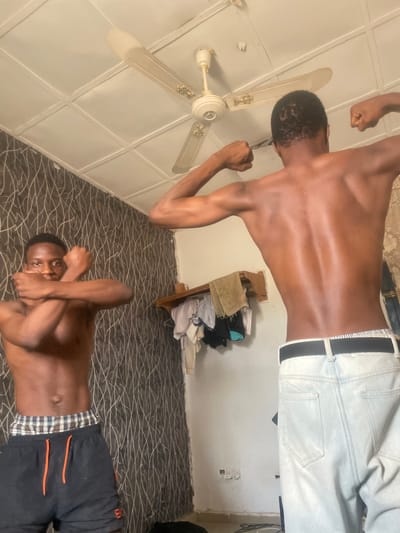 This image, captured indoors in Bauchi, Nigeria, features two young, dark-skinned males posing. The photograph is rotated 90 degrees clockwise from its intended orientation.
One male is positioned as if standing, shirtless and wearing light-colored pants, flexing his biceps with both arms raised. His back is largely towards the viewer, with his head slightly turned. Above him, a white ceiling fan with four blades and golden accents is mounted on a white, panelled ceiling.
The second male is lying on a dark floor covered with a patterned carpet or rug featuring wavy, light-colored lines. He is also shirtless, wearing dark shorts with orange stripes and a plaid waistband. He looks directly at the camera with his arms crossed over his chest in a casual, possibly playful, pose.
In the background, a light-colored wall is visible, against which stands a wooden shelf or small piece of furniture laden with folded clothes. A white electrical outlet is also discernible on the lower part of the wall. The scene suggests a relaxed, candid, or playful moment taking place within a personal indoor setting, likely a room in a home. No text is visible in the image.