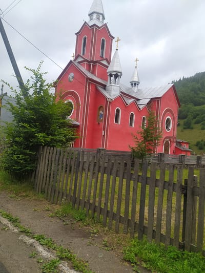 The photograph captures a vibrant red church with silver-colored spires topped with golden crosses, located in Luhy, Ukraine. The church exhibits distinct architectural features, including arched windows, a circular window on the side, and decorative white trim. A fresco of a religious figure is visible on the facade. The church is surrounded by lush green trees and vegetation, with a backdrop of rolling, forested hills under an overcast sky. A weathered wooden fence runs along the foreground, separating the churchyard from a dirt path and a small roadside area with scattered weeds. A utility pole with wires is also present on the left side of the image. The scene depicts a peaceful, rural religious establishment.
