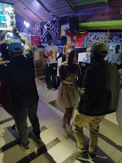 The image captures an indoor scene, likely a bar, lounge, or entertainment venue, characterized by dim lighting and vibrant accent colors, suggesting an evening or nighttime setting. The perspective is from behind and slightly to the side of several individuals who are standing and socializing.

In the foreground, a person is seen from the back, wearing a dark top and dark pants, with a light blue baseball cap worn backward. Their attention seems to be directed towards a screen or monitor visible in the lower-left corner of the frame, which displays a bright, possibly animated or sports-related image.

Further into the mid-ground, a woman with braided hair is visible, turned slightly to her left, wearing a light-colored, possibly patterned dress or top. She appears to be holding a smartphone in her hand. To her right and slightly behind her stands another person, seen from the back, wearing a dark jacket, a baseball cap, and light brown or khaki pants, with dark shoes.

In the background, several other individuals can be seen. One person in a bright red hoodie is visible, holding up a smartphone, possibly taking a photo or video. Another person in a white t-shirt with a graphic design is partially visible behind them. The background features decorative elements, including white, intricately cut-out panels on the wall, and shelving units. Colored lighting, including blue, green, and purple hues, illuminates different parts of the space, casting a lively glow.

The floor is tiled with alternating light-colored and dark-patterned squares, adding to the decorative interior. The overall impression is one of a casual gathering where people are relaxed and engaged in conversation or observing activities around them. No discernible text is visible in the image.
