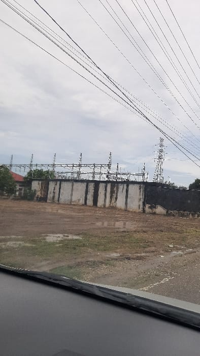 A view from inside a vehicle shows an electrical substation with tall power line structures along the left. A long concrete wall and fenced area protect the equipment. Overhead power lines extend far into the distance. Patches of dry grass and brown earth cover the ground next to the station. The sky is overcast. The vehicle's interior is visible on the right.