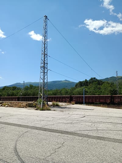 The image depicts an outdoor scene at a railway yard in Zlatitsa, Bulgaria. In the foreground, a paved area with visible tire marks and cracks suggests recent vehicle activity or wear and tear. A tall, metal lattice tower, likely an electrical pylon for the railway, dominates the left side of the frame. Overhead, taut wires extend from the tower, disappearing into the distance and implying an active power supply for trains.

In the mid-ground, a long train composed of numerous dark brown, open-top freight cars is parked on railway tracks. These cars are laden with what appears to be rocky material or ballast. Behind the train, a dense line of lush green trees forms a natural barrier, with rolling, verdant hills rising further in the background. The sky is a clear, bright blue, dotted with a few scattered white clouds. A small, dark object, possibly an airplane or a bird, is visible in the upper right quadrant of the sky. The overall impression is one of a functional, industrial railway environment set against a backdrop of natural, hilly terrain.