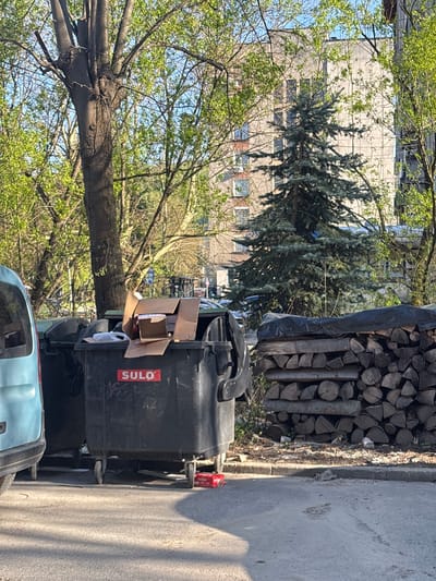 This outdoor scene, captured under bright daylight in what appears to be Troyan, Bulgaria, depicts a side street or alleyway lined with everyday objects and vegetation. The image is oriented vertically, showing elements from foreground to background.
In the immediate foreground, the top portion of a light blue car is visible at the bottom left. Adjacent to it are two large, black, wheeled garbage bins. The bin closer to the viewer has an open cardboard box resting on its lid, with another cardboard box visible inside. The brand name "SULO" is clearly visible in red text on the side of this bin, along with some faint, illegible white markings below it.
Behind the bins, a large, neatly stacked pile of cut firewood is present, partially covered by a dark, possibly blue or black, tarp. This stack is situated along a concrete curb that separates the asphalt surface from a patch of ground.
The midground and background are characterized by abundant greenery and a building. Several deciduous trees with bright green leaves, indicative of spring or early summer, frame the left and upper parts of the scene. A darker green coniferous tree, possibly a spruce or pine, stands prominently in the center-left. Behind the trees, a multi-story building with a light-colored facade and multiple windows is visible, suggesting a residential or commercial structure. The sky above is a clear, bright blue.
The strong shadows cast by the objects suggest a sunny day, likely mid-morning or afternoon. No people are visible, but the presence of the garbage bins and firewood indicates human activity in the area.