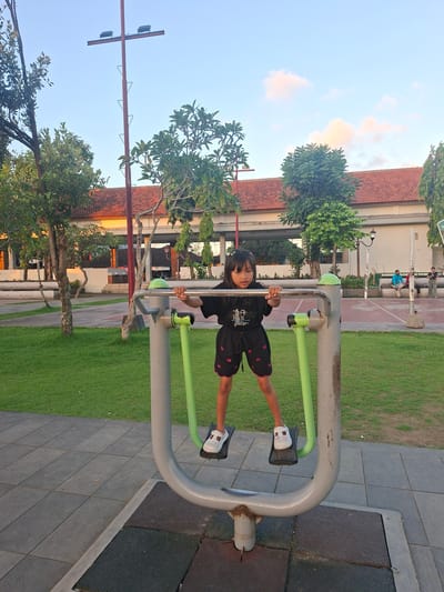 In Gianyar, Indonesia, a young girl is photographed outdoors at a public park. She stands on the footplates of a grey and green outdoor exercise machine, holding onto its handlebars. The girl wears a black t-shirt and black shorts with a red trim and embroidered pattern. She has dark hair and is wearing white sandals. The exercise equipment is situated on a paved area surrounded by a manicured lawn and trees. In the background, a building with a red-tiled roof is visible, along with a tall lamppost. The sky above is clear with a few light clouds.