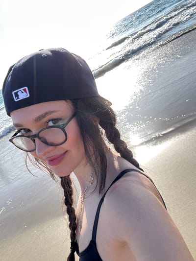 The image displays a close-up selfie of a female individual on the beach in Manhattan Beach, United States. The subject is wearing a dark blue baseball cap with a white and red MLB logo on its left side, tortoise-shell framed glasses, and a black swimsuit top. Her dark brown hair is styled in two braids, visible over her shoulders, and she wears a thin, silver-colored beaded necklace with a small charm. Her lips are tinted pink, and she is looking towards the camera with a slight smile. The background shows the ocean with small waves and the wet sandy shoreline, heavily illuminated by bright sunlight causing significant glare and reflections on the water and sand.