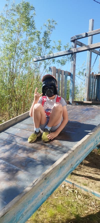 A young child, estimated between six and ten years old, is seated cross-legged on a weathered, dark blue metallic slide or ramp, part of a dilapidated wooden playground structure in Zhatay, Russia. The child is wearing a black Darth Vader mask, a grey t-shirt with "SURF" visible, grey shorts, light blue socks, and bright green athletic shoes. A red baseball cap is worn backward beneath the mask, and red backpack straps are visible over the child's shoulders. The child's left hand is raised, performing a "rock on" hand gesture. To the left of the child, dense green foliage with tall, slender branches extends upward. To the right, a worn, blue-painted wooden structure, possibly a playhouse, stands against a clear, bright blue sky. The ground visible at the bottom right is dry earth with sparse green grass. Strong sunlight illuminates the scene, casting distinct shadows.