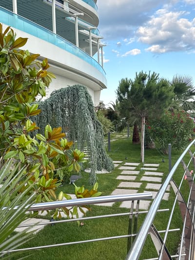 The image depicts a lush garden area adjacent to a modern building with a curved glass facade. The garden is meticulously maintained with vibrant green grass and a pathway made of square stone pavers. Several trees and plants are visible, including a tall pine tree, a weeping conifer, and broad-leafed evergreen shrubs. A sleek, metallic railing runs along a curved structure, possibly a balcony or walkway, adding a contemporary touch to the scene. In the background, a bright blue sky with scattered white clouds suggests fair weather during the daytime. The location appears to be a well-landscaped outdoor space, potentially a residential or commercial property, with a view of the sea or a large body of water implied by the architecture and greenery. There is no visible text in the image. The overall impression is one of tranquility and modern design integrated with nature.