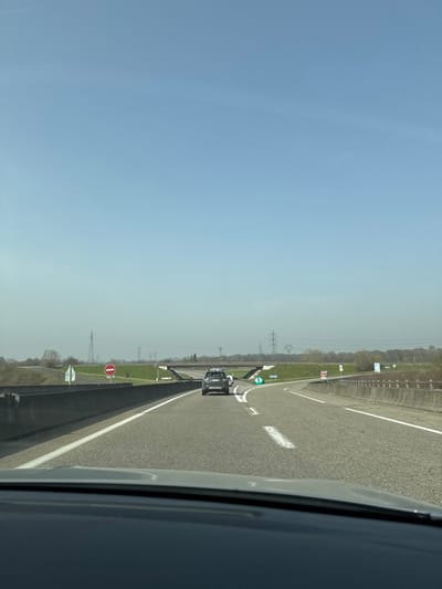 The image depicts a highway scene near an exit ramp junction in Eschau, France, viewed from inside a vehicle looking forward over the dashboard. The sky is clear and light blue, indicating fair weather. The asphalt roadway features white dashed and solid lane markings. Ahead, a dark-colored SUV is positioned in the right-hand lane of the main thoroughfare. A second, lighter-colored vehicle is partially visible behind and to the left of the SUV, suggesting movement on the road or an adjacent lane.
To the right, an exit ramp curves away from the main highway, marked by a green chevron sign indicating a right turn. Concrete barriers line both sides of the highway. On the left side of the road, a red circular "No Entry" sign with a white horizontal bar is prominently displayed on a grassy verge. In the mid-ground, a concrete overpass spans across the highway. The distant horizon reveals several metal lattice transmission towers against the sky, with sparse deciduous vegetation bordering the highway. No individuals are visible in the scene.