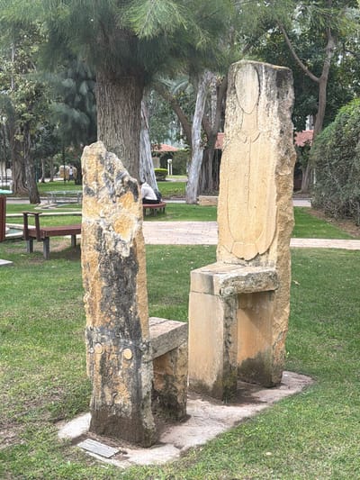 This image depicts a stone monument in what appears to be a park-like setting in Herzliya, Israel. The monument consists of two large stone pieces, seemingly carved with symbolic designs. The upper stone features a carved, oval shape and intricate detailing. The lower stone, darker in tone, has a rougher texture. The monument is set on a stone base surrounded by green grass. Trees are visible in the background, along with benches and a path. The overall environment suggests a peaceful public space within the city of Herzliya.