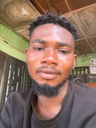 A close-up headshot shows a Black man with short, dark, curly hair and a dark beard. He is wearing a black shirt. The background consists of a wooden wall painted in a faded beige and green, with a window frame on the right and a calendar hanging on the wall. The location is likely Chikaji, Nigeria.