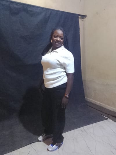 An indoor, full-body portrait depicts an adult woman standing and posing for the camera. She has dark skin and her braided hair falls over her shoulders. She wears a light-colored (off-white or light beige) short-sleeved polo shirt with a textured pattern, black trousers, and light-colored sneakers with blue accents. A watch with an orange-brown band is visible on her left wrist, and she has gold-colored earrings. The woman faces forward, with her body slightly turned, looking directly at the camera with a subtle, pleasant smile.
The setting appears to be a prepared photographic area. A dark grey or black fabric backdrop is hung behind her, covering the majority of the background. To the right, a light brown or tan wall is partially visible. The floor is light-colored marble or tiled with dark grey veins. The lighting suggests an indoor environment. The image is taken in Kuru, Nigeria. No visible text is present.