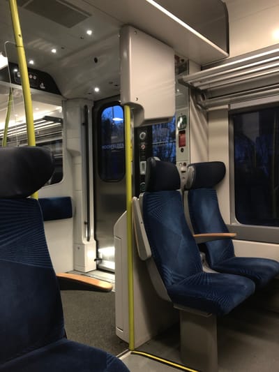 The image shows the interior of a train car. Blue, patterned seats are arranged in pairs facing each other, with a small table between them. Windows show an outdoor scene blurred from motion. Above the seating area, the ceiling has lighting panels and a ventilation system. The walls are white and sleek, with controls and displays visible.