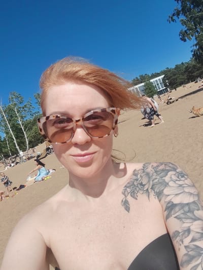 This is a selfie taken by a fair-skinned woman with red hair and freckles, wearing large, tortoise-shell patterned sunglasses. She has a floral tattoo on her left arm and shoulder. She is on a sandy beach under a clear blue sky. In the background, there are people sunbathing and walking. There are also trees and a modern white building visible. The woman appears to be in Solnechnoye, Russia, likely during a sunny day.