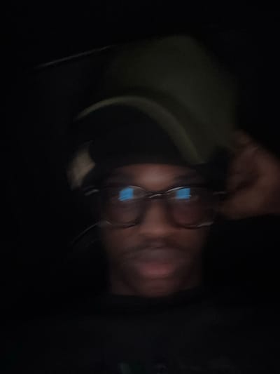 The image is a dimly lit, blurry selfie of a person wearing glasses and a hat. The person appears to be of African descent, with dark skin and hair. They are looking directly at the camera, and their mouth is slightly open. The background is dark and indistinct, making it difficult to determine the location. However, the question specifies Enugu, Nigeria, and while not evident from the image itself, if we assume that context, this could be a personal photo taken in a private residence or vehicle within the city. The lighting suggests it might be nighttime or an indoor setting with minimal light. The quality of the image, with its blurriness and low light, indicates it was likely taken with a mobile phone camera under suboptimal conditions. The focus is on the person's face, with the glasses reflecting some light, possibly from a screen or ambient light source. The hand of the person is visible on the right side of the image, adjusting the hat.
