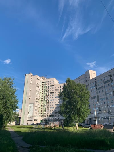 This is a street-level, eye-level, medium shot of an apartment complex in Severodvinsk, Russia, on a clear, sunny day. The building dominates the frame, a long, multi-story concrete structure with numerous windows and balconies. To the left, a lush green tree stands tall, its leaves full and vibrant. In the foreground, a concrete path winds through an area of tall grass. In the middle ground, a playground with colorful equipment is visible. A few cars are parked near the building, and a large, rusted dumpster sits to the right. The sky above is a brilliant blue, with a few wispy white clouds scattered across it. The overall impression is one of an ordinary urban scene, bathed in the bright light of a summer day.