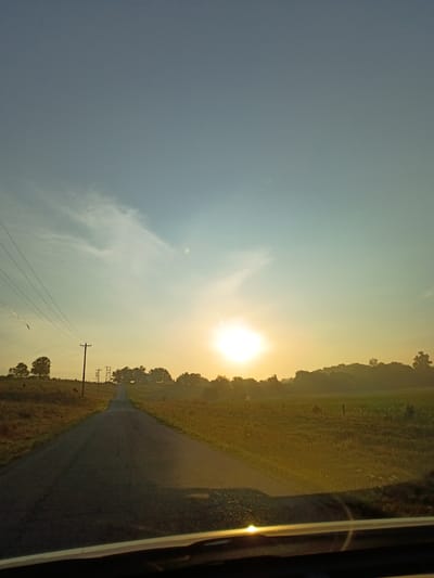 The image depicts a rural road in Camargo, United States of America, at sunrise. The sun is low on the horizon, casting a warm glow and creating a hazy atmosphere. The road stretches into the distance, flanked by fields on either side. Power lines run along the left side of the road, supported by utility poles. In the background, a line of trees and rolling hills can be seen. The foreground shows the dashboard of a vehicle, suggesting the image was taken from a car. The overall scene evokes a sense of peace and tranquility, characteristic of a rural landscape at the beginning of a new day.