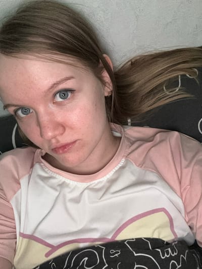 The image presents a close-up portrait of a single female individual in an indoor environment within Nizhnekamsk, Russia. The subject has fair skin with a slight reddish tone, light blue eyes, and light brown hair. She looks directly into the camera with a neutral to slightly somber expression. Her hair is partially visible, extending towards the right side of the frame, and a small gold-colored hoop earring is present in her right earlobe. She is wearing a long-sleeved top with a white torso and light pink raglan sleeves; the white section displays a light yellow and pink graphic design near the lower edge. The individual is positioned in a reclining manner. A dark grey fabric with white abstract patterns is visible around her, functioning as both a bedding item and a background element. Behind her head, a textured, light grey wall is visible. No other individuals, significant objects, or specific actions beyond posing for the photograph are discernible.