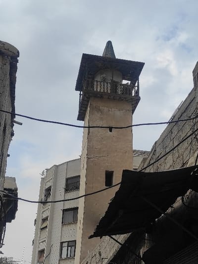 The image displays an urban scene within Al-Qanawat Municipality, Syria. A weathered, light-brown minaret with a square shaft and a single rectangular opening dominates the central view. The minaret's summit features a wooden balcony with a balustrade, topped by a dark, pointed, pyramidal roof. Two loudspeakers are discernible within the balcony structure. To the left and rear of the minaret, a taller, modern, light-colored multi-story building with multiple dark-framed windows and balconies is visible. On the right side, dark, tattered fabric shades are suspended above a structure, partially obscuring a rough-textured wall. A damaged wall segment is present on the far left. Multiple black electrical or communication wires crisscross the frame against an overcast sky with visible clouds. No individuals or specific actions are depicted.